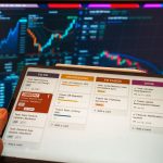 Tablet exibindo um aplicativo de gerenciamento de tarefas diante de monitores com gráficos de negócios.