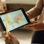 Mãos seguram um tablet que exibe um mapa-múndi, representando o uso de tecnologia digital na educação em escala global.