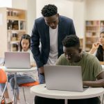 Estudantes usam laptops em uma sala de aula enquanto recebem orientação de um professor.