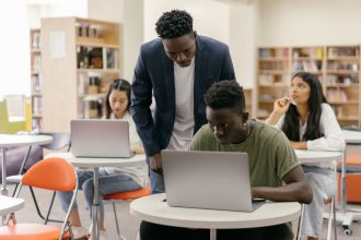 Estudantes usam laptops em uma sala de aula enquanto recebem orientação de um professor.