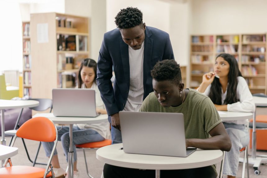 Estudantes usam laptops em uma sala de aula enquanto recebem orientação de um professor.