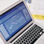 Laptop exibindo gráficos e análises financeiras sobre uma mesa, cercado por documentos.