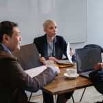 Profissionais de diferentes perfis discutem um plano de negócios em torno de uma mesa, com laptop e documentos.