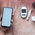 Mão segura um celular ao lado de um glicosímetro e outros materiais médicos sobre uma superfície de madeira.