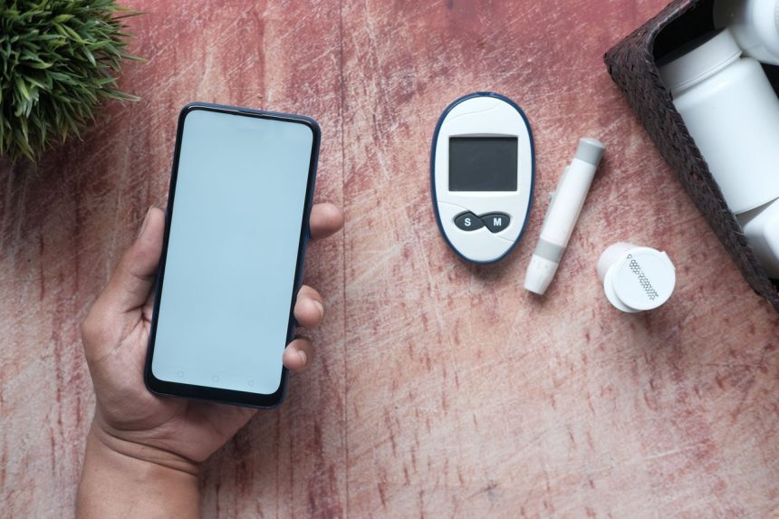 Mão segura um celular ao lado de um glicosímetro e outros materiais médicos sobre uma superfície de madeira.