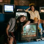 Duas mulheres jogam em consoles retrô diante de televisores antigos em um ambiente interno.