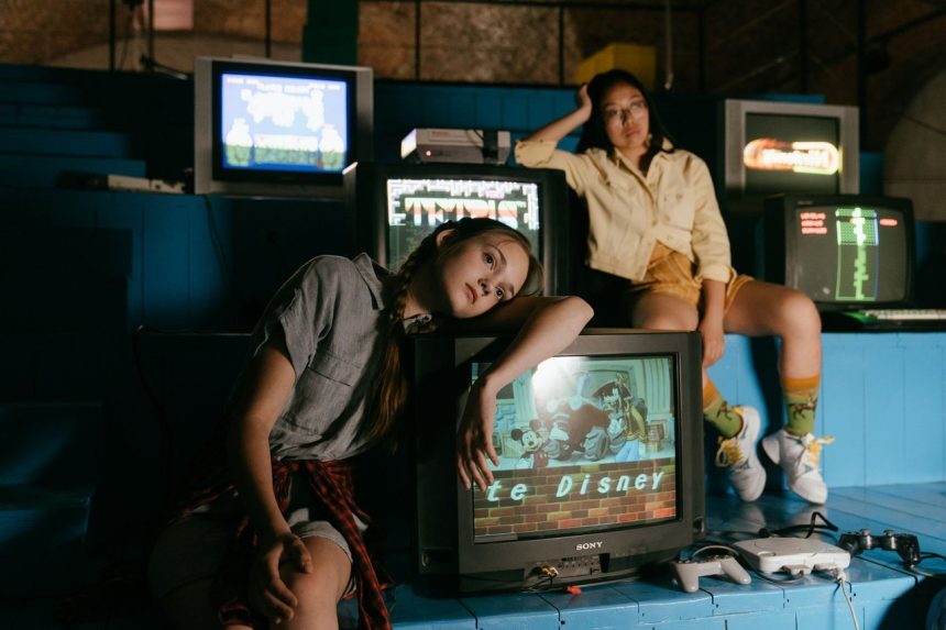 Duas mulheres jogam em consoles retrô diante de televisores antigos em um ambiente interno.