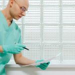 Profissional da saúde, usando luvas e roupa hospitalar, examina atentamente um documento.