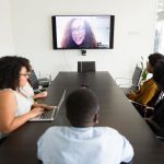 Equipe reunida em uma mesa de conferência participa de uma chamada de vídeo com um colega exibido em uma televisão.