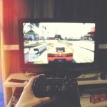 Mão segurando um controle de videogame diante de uma televisão.